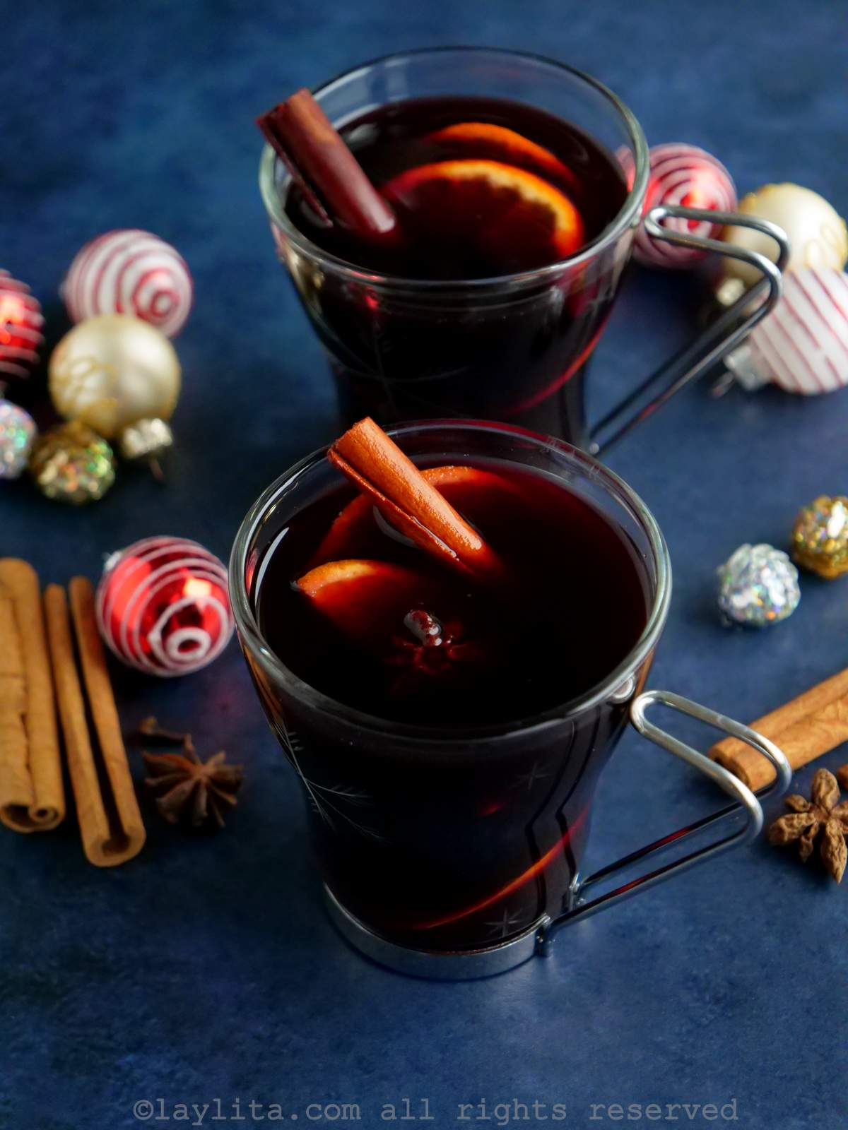 Dos vasos con vino caliente en una superficie azulada con decoraciones navideñas y especias al lado