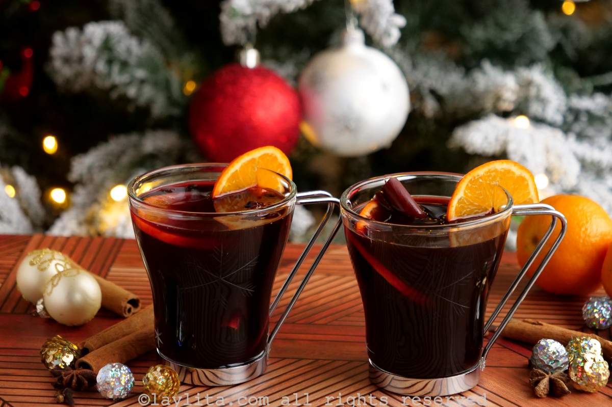 Dos copas de vidrio con vino caliente adornadas con rodajas de naranja y astas de canela con un árbol de Navidad de fondo.