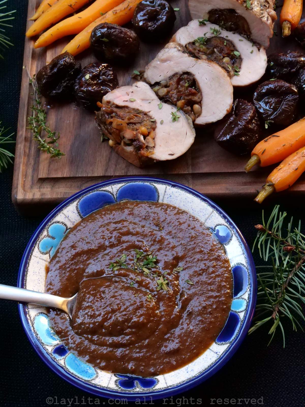 Un bol azulado lleno de salsa espesa de ciruelas, en el fondo hay rodajas de cerdo relleno, ciruelas enteras y zanahorias asadas