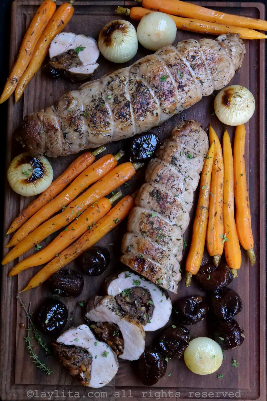 Una tabla de madera con verduras asadas y dos solomillos de cerdo, el uno tiene algunas rodajas cortadas mostrando el relleno de ciruelas