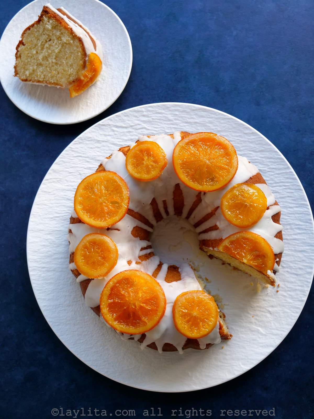 Una torta de naranja decorada con glaseado y rodajas de naranja confitadas en un plato blanco en una superficie azulada con una rebanada de la torta al lado