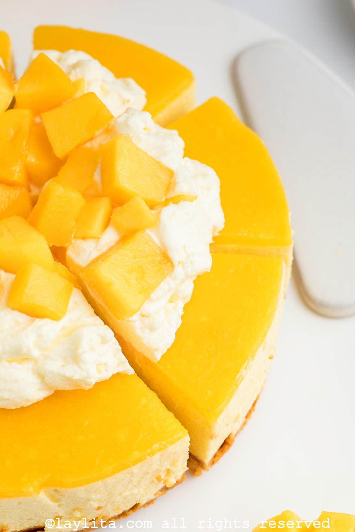 Primer plano de una tarta de queso o un cheesecake con mango, con algunos trozos cortados en rodajas, crema batida y mango en cubos encima.