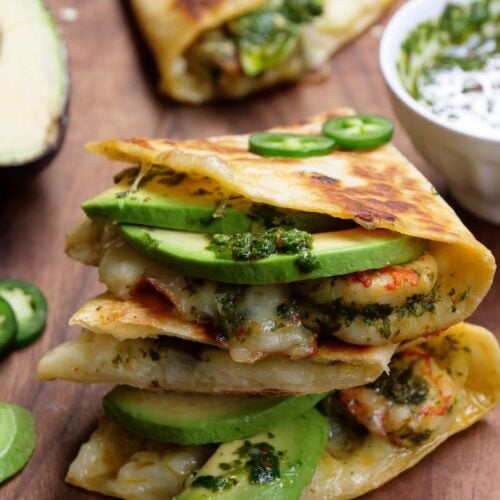 Dos quesadillas de camarones apiladas una encima de la otra sobre una tabla de madera junto a un bol de chimichurri.