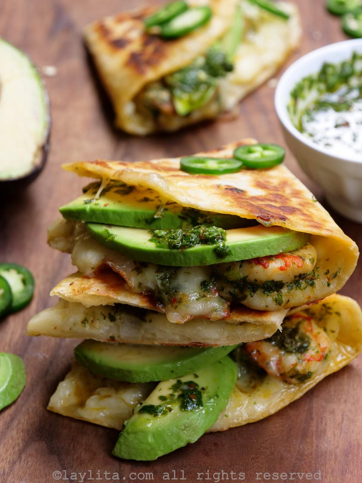 Dos quesadillas de camarones apiladas una encima de la otra sobre una tabla de madera junto a un bol de chimichurri.