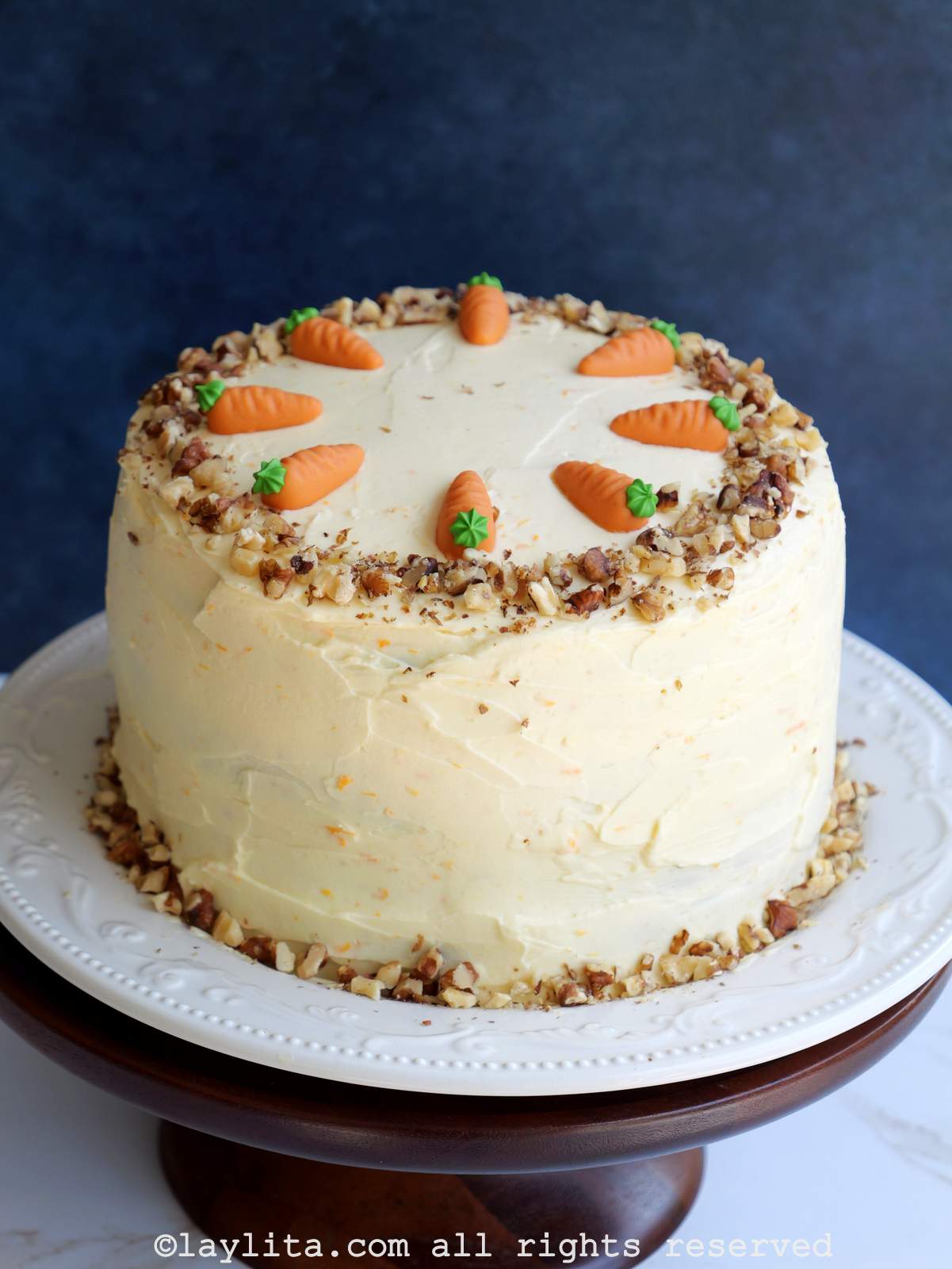 Un pastel de zanahoria decorado con nueces y zanahorias decorativas en miniatura en un plato blanco encima de un pedestal para pasteles