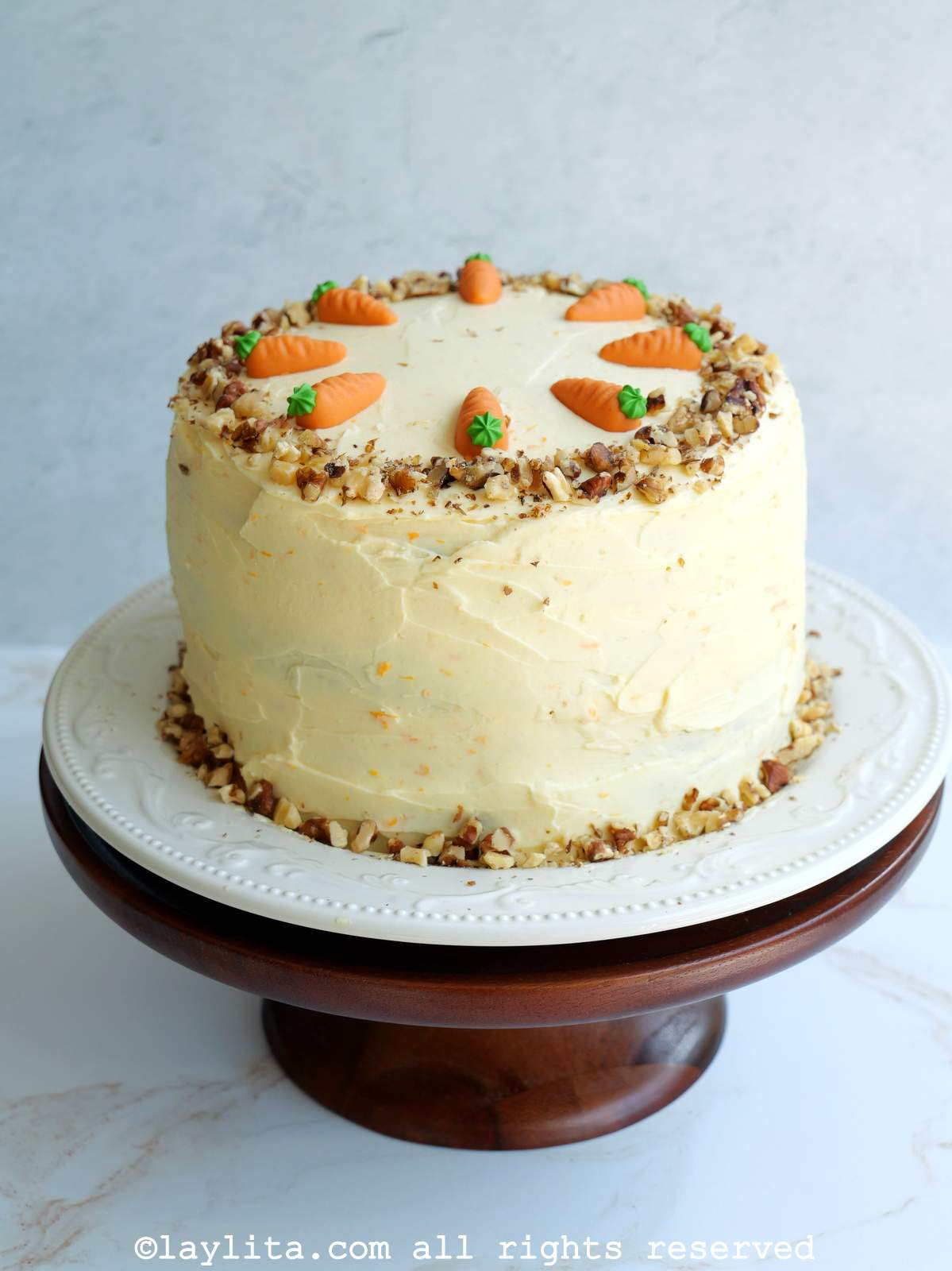 Un pastel de zanahoria decorado con nueces y zanahorias decorativas en miniatura en un plato blanco encima de un pedestal para pasteles