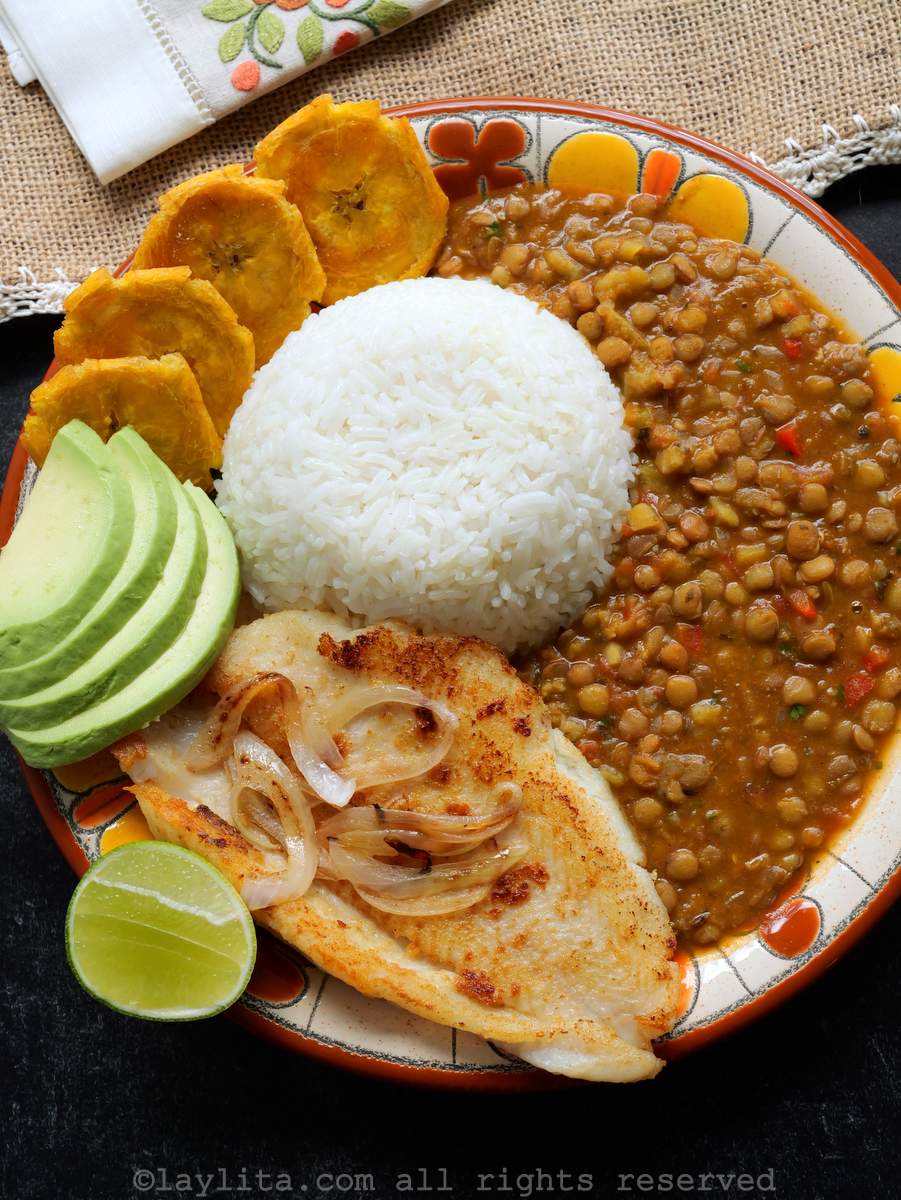 Vista superior de un plato servido con arroz, lentejas, pescado a la plancha, aguacate, plátanos fritos y cebollas.