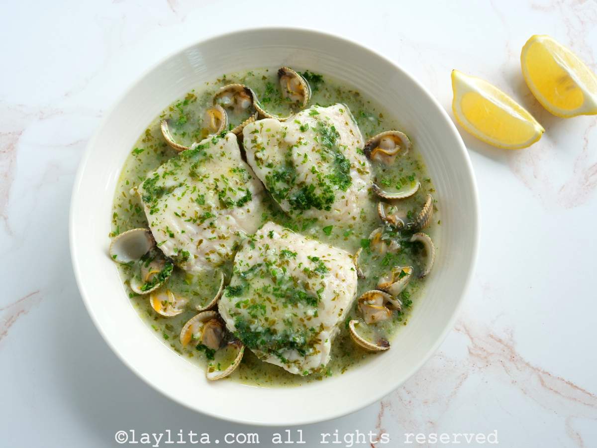 Un plato sopero lleno de pescado en salsa verde con almejas y dos rodajas de limón al lado.