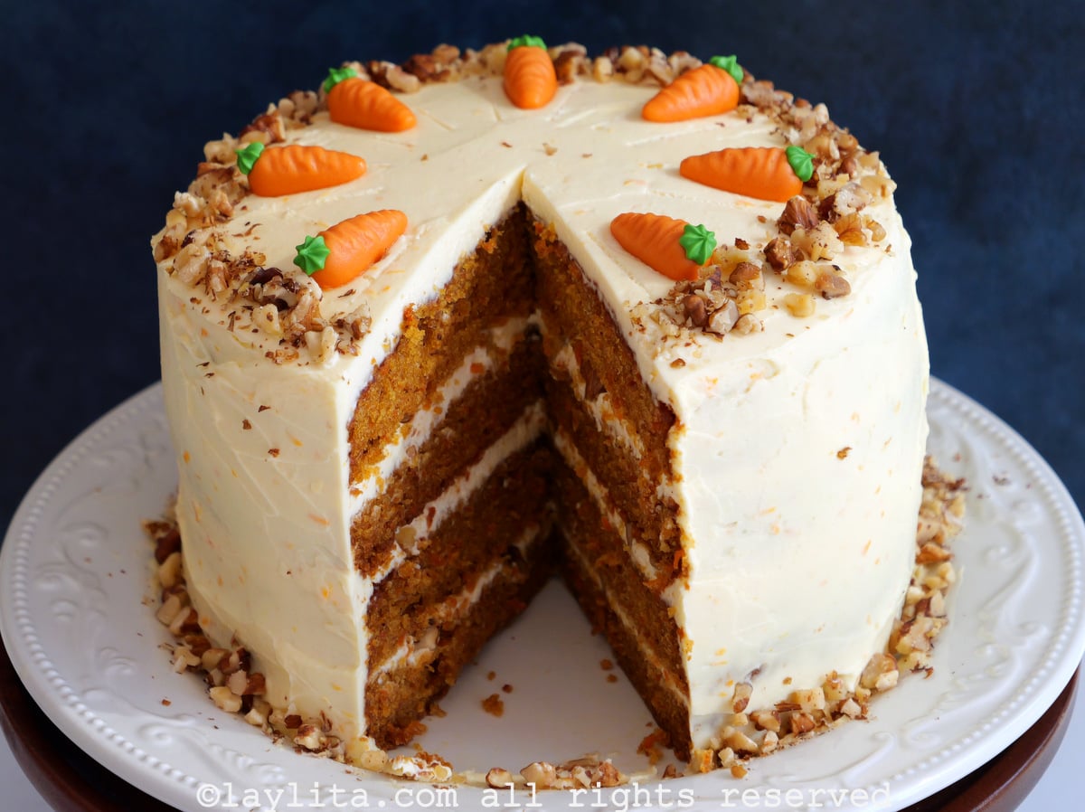 Un pastel de zanahoria con capas de queso crema y nueces por encima, al pastel le falta una rebanada