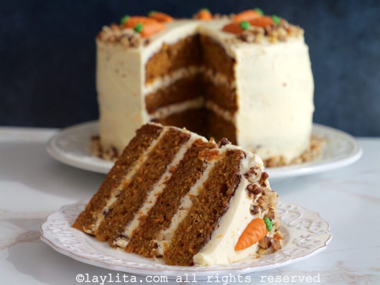 Una rebanada de pastel de zanahoria en capas sobre un plato blanco, con el resto del pastel en la parte posterior