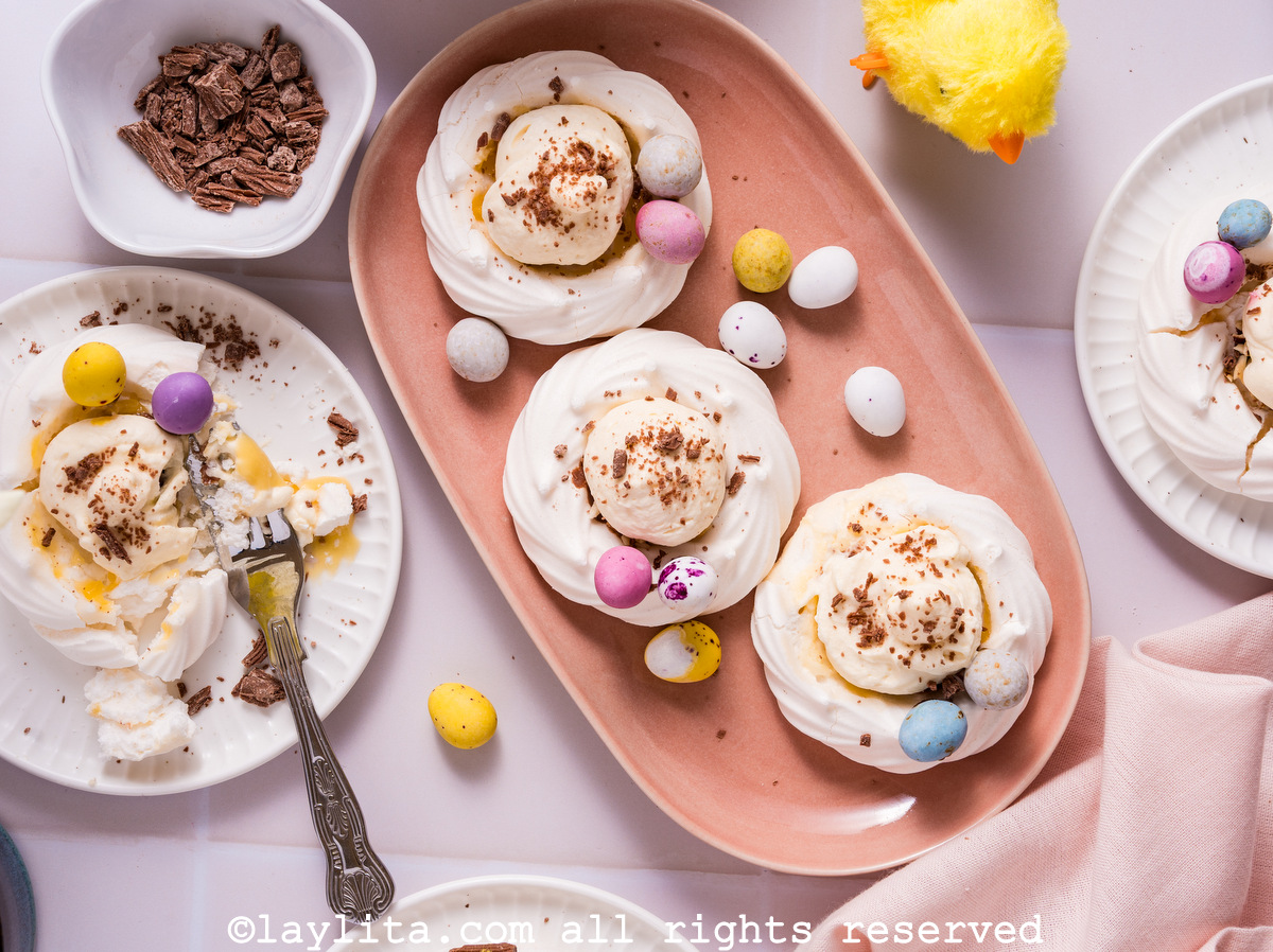 Una bandeja con tres mini pavlovas o nidos de merengue rellenos para Pascua con decoraciones de Pascua alrededor
