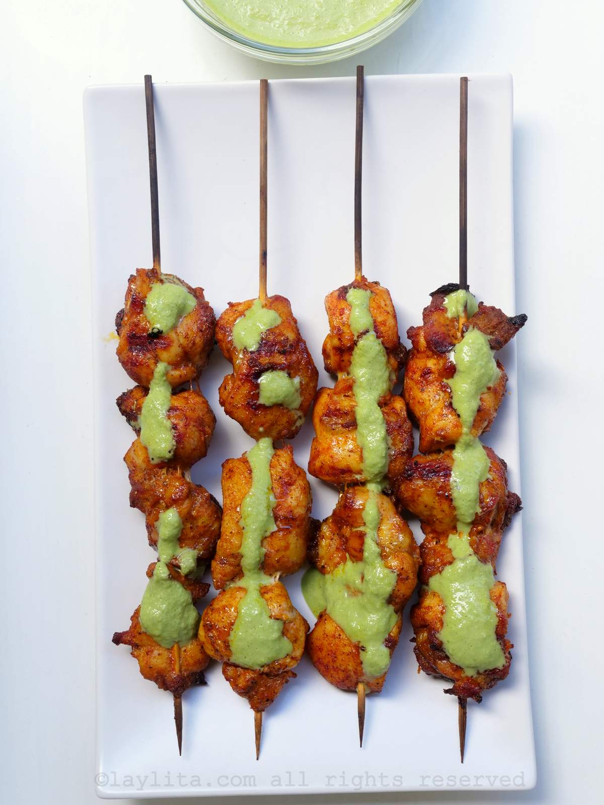Cuatro brochetas de pollo en un plato blanco con salsa verde encima