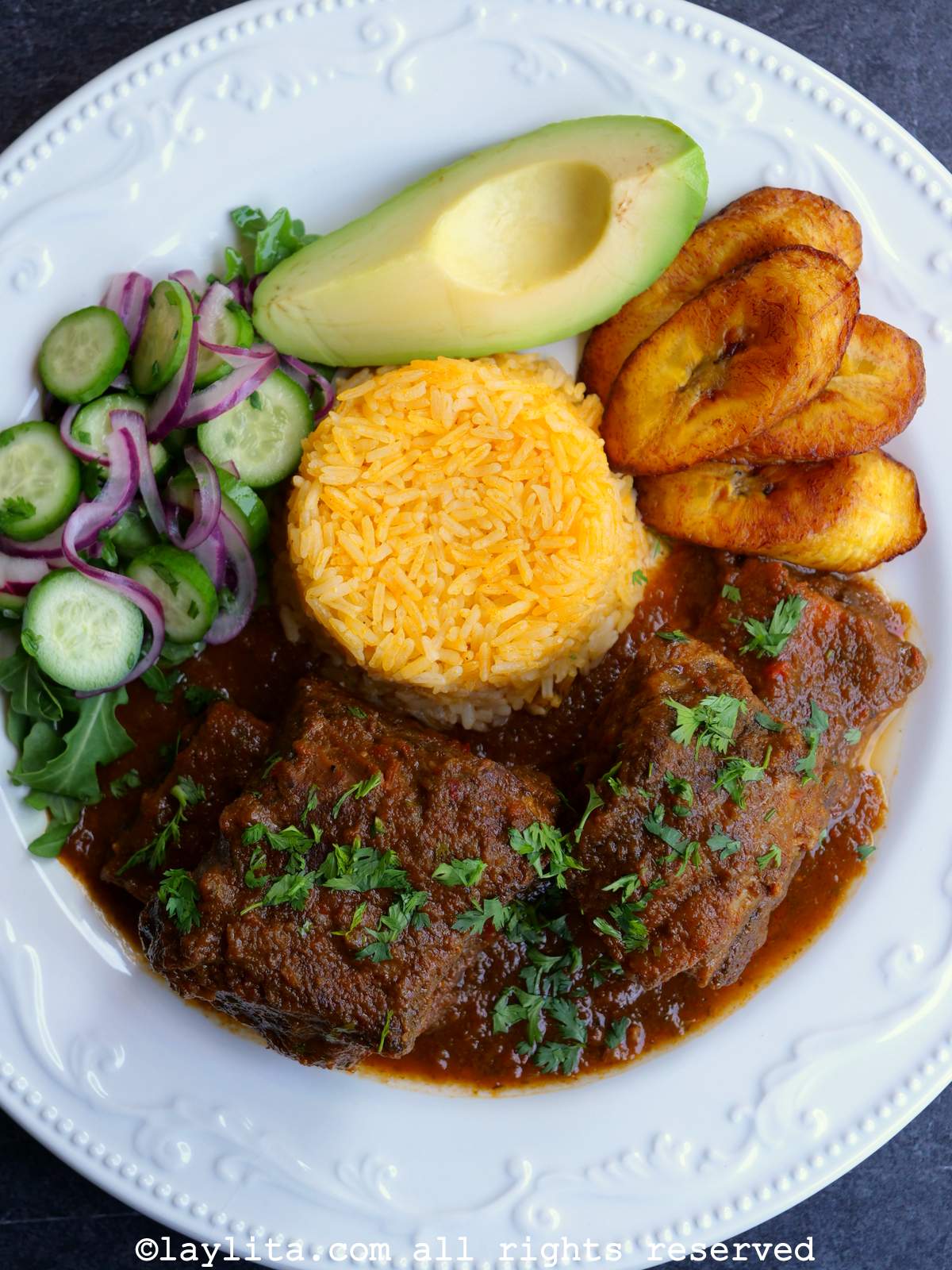 Un plato blanco con seco de costilla de res, arroz amarillo, plátanos maduros fritos, aguacate y ensalada de pepino.
