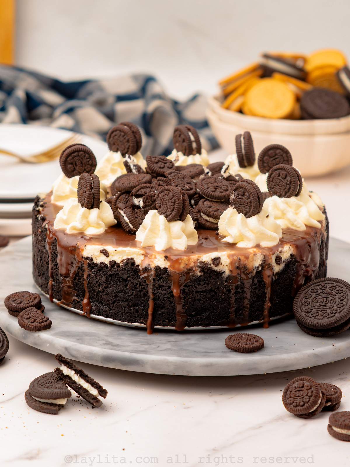 Cheesecake de Oreo Sin Horno con cobertura de salsa de chocolate, crema batida y galletas adicionales, en una bandeja sobre la encimera de la cocina.