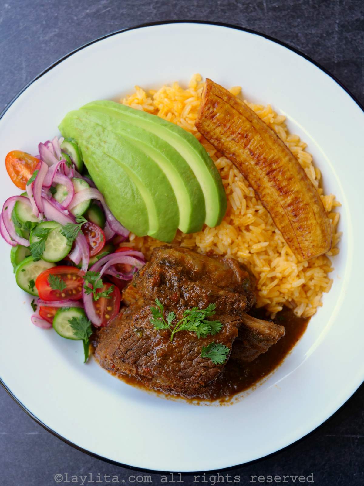 Vista superior de un plato blanco con seco de costillas de res, arroz amarillo, rodajas de aguacate, plátano frito y ensalada.
