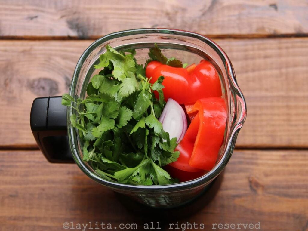 Una licuadora con tomate, pimientos, cebolla y hierbas verdes