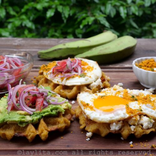 Tres waffles de plátano verde macho con guarniciones como puré de aguacate, huevo frito, curtido de cebolla, encima de una tabla de madera