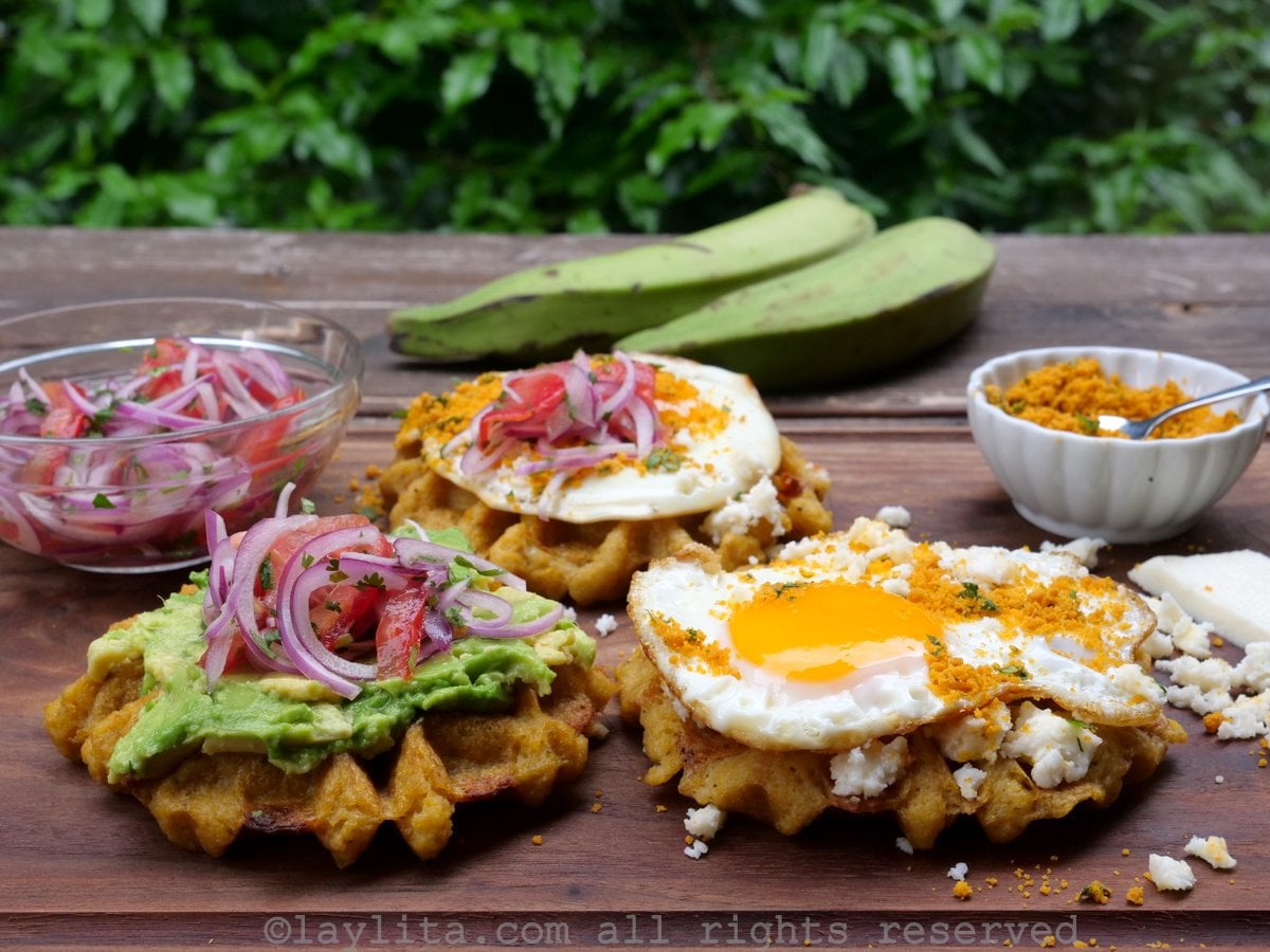 Tres waffles de plátano verde macho con guarniciones como puré de aguacate, huevo frito, curtido de cebolla, encima de una tabla de madera