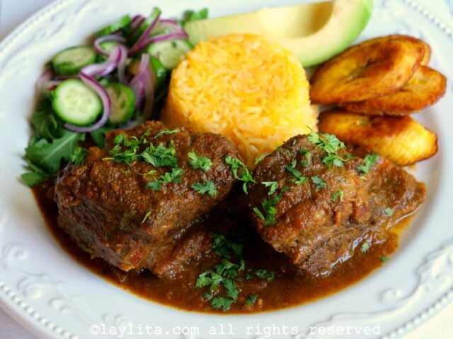 Seco de costilla de res en un plato blanco acompañado con arroz amarillo, plátanos maduros fritos, aguacate, y ensalada de pepino