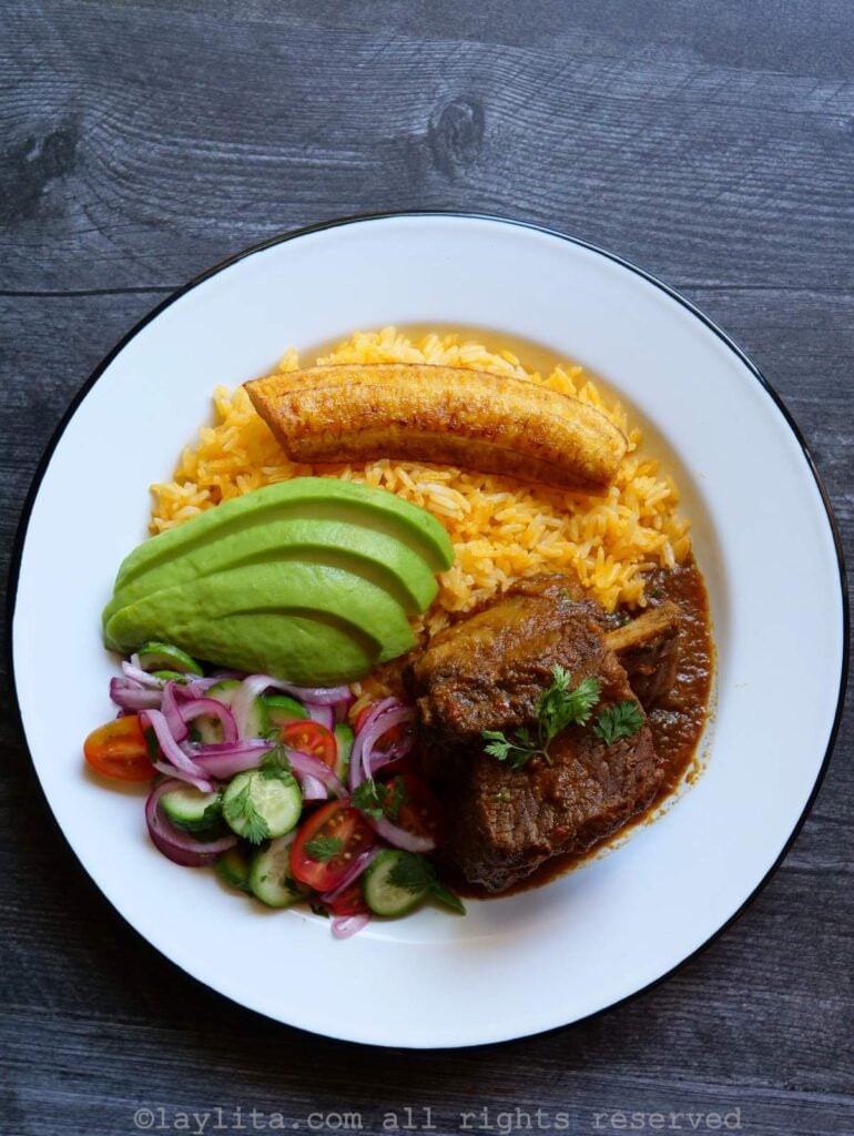 Vista superior del seco de costilla guisado, arroz amarillo, plátanos maduros fritos, aguacate y ensalada en un plato blanco con borde oscuro sobre una mesa de madera oscura.