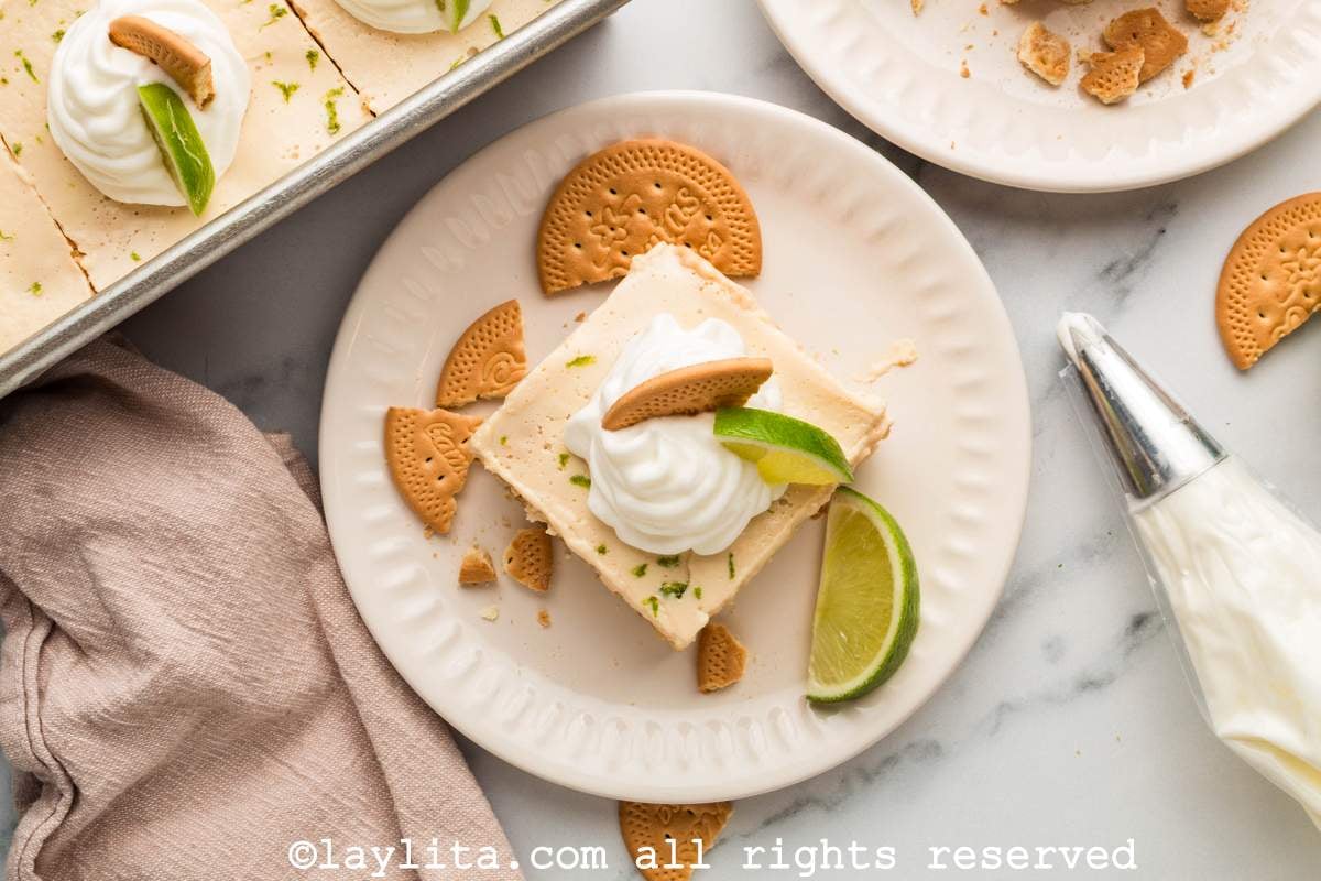 Vista superior de un plato con un pedazo de carlota de limón decorado con crema chantilly, al lado se ve una bolsa con la crema y el resto del postre en una bandeja
