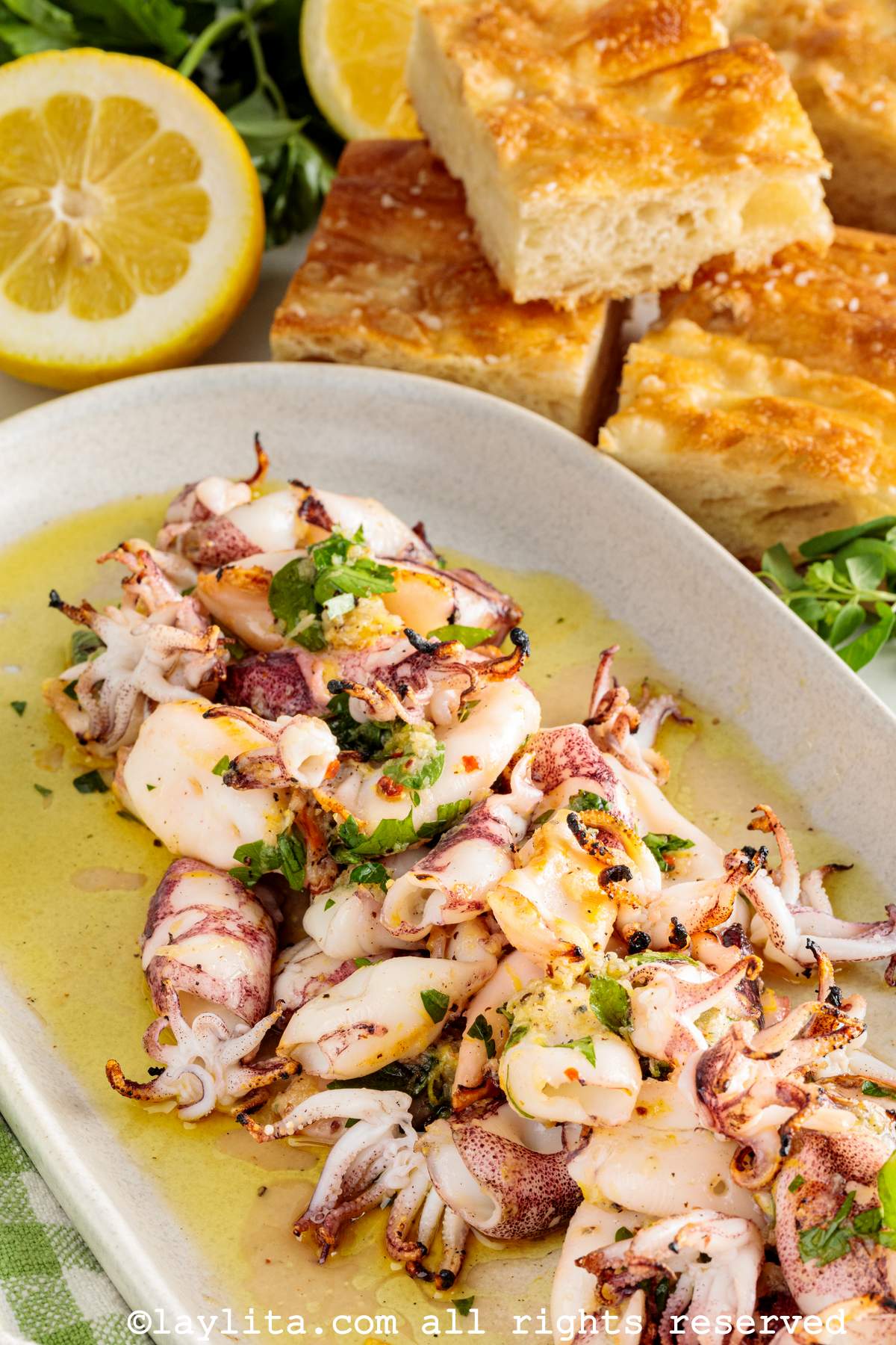 Una bandeja con ensalada de calamar asado con trozos de pan focaccia y limones en el fondo