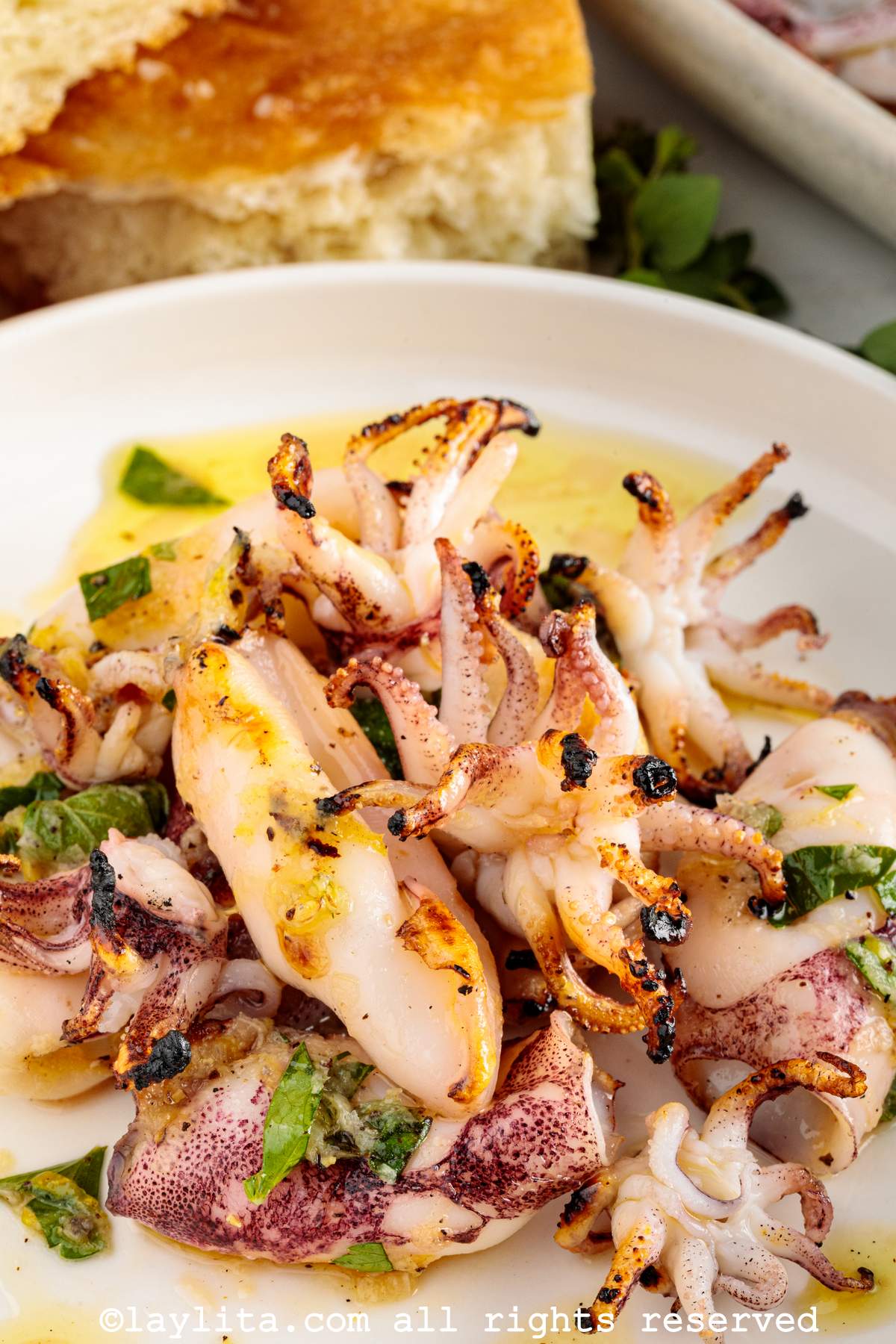Primer plano de calamares asados en un plato con aderezo de limón y hierbas, con trozos de pan en el fondo