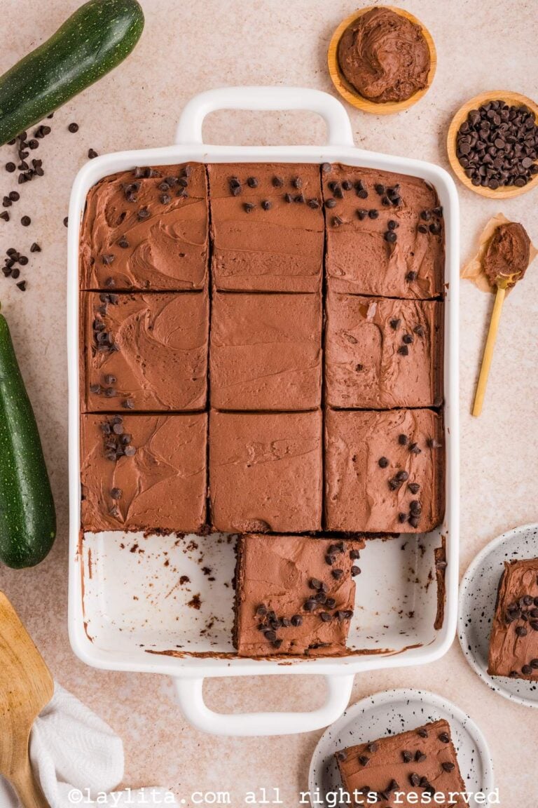 Un molde blanco con un pastel de chocolate y zucchini decorado con betún y chispas de chocolate, con dos rebanadas faltantes