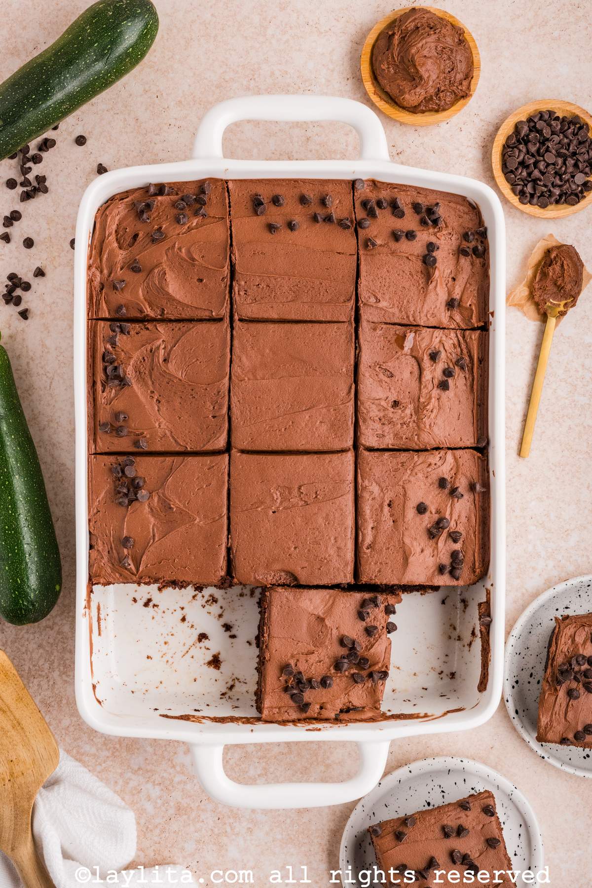 Un molde blanco con un pastel de chocolate y zucchini decorado con betún y chispas de chocolate, con dos rebanadas faltantes