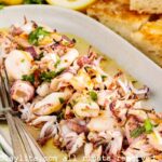Una ensalada de calamares con limón y perejil con utensilios de servir al lado y trozos de pan en el fondo