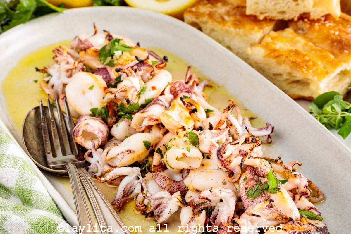 Una ensalada de calamares con limón y perejil con utensilios de servir al lado y trozos de pan en el fondo