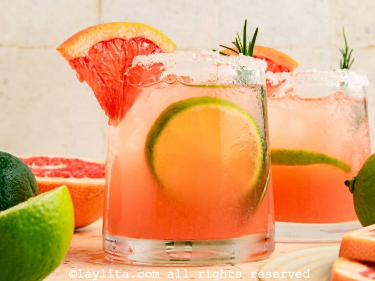 Dos vasos del cóctel Paloma, decorados con escarcha de sal y rodajas de pomelo y limón con frutas cítricas adicionales alrededor de los vasos