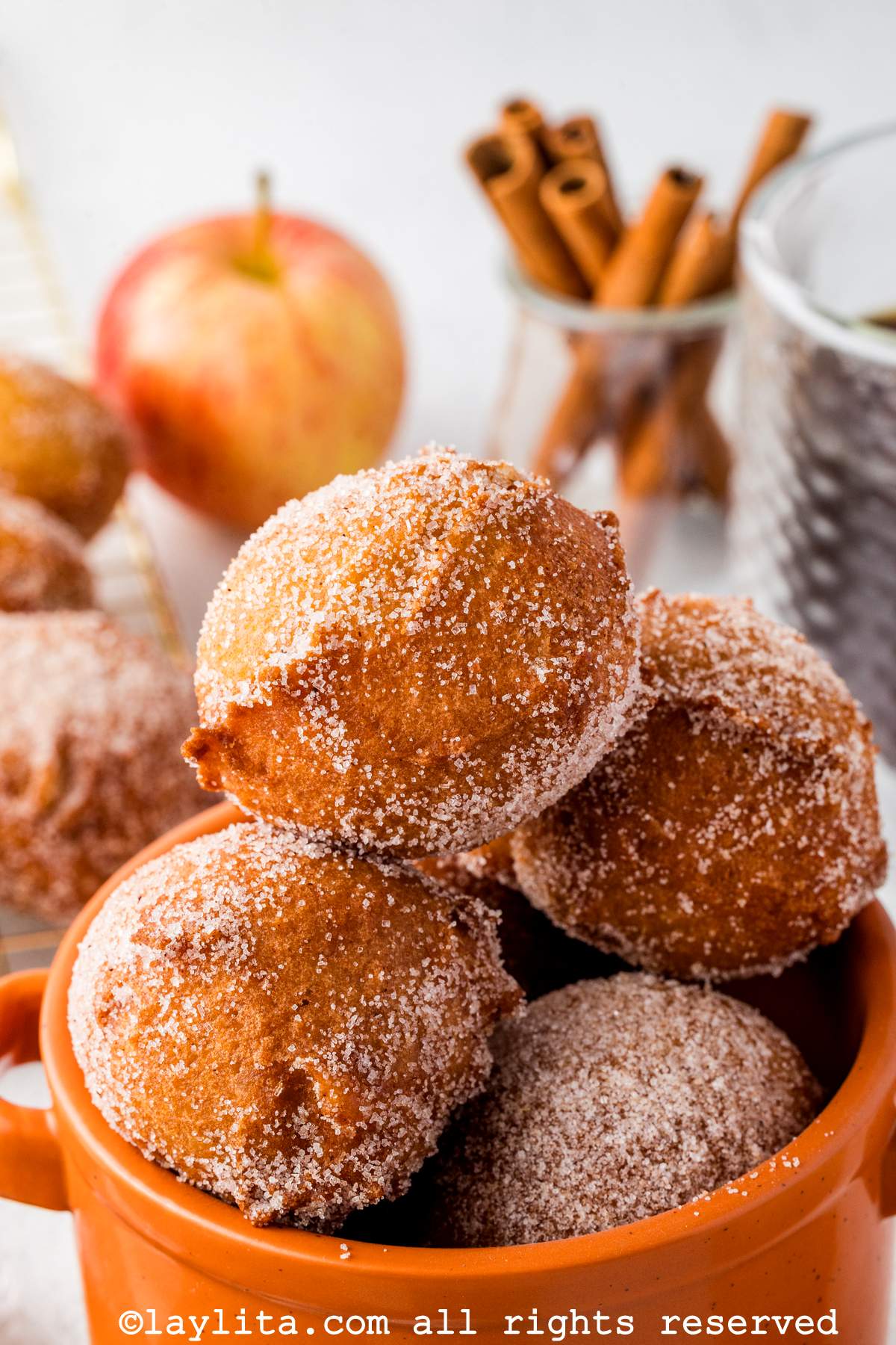Varios buñuelos de manzana en un tazón anaranjado con astas de canela y una manzana en el fondo