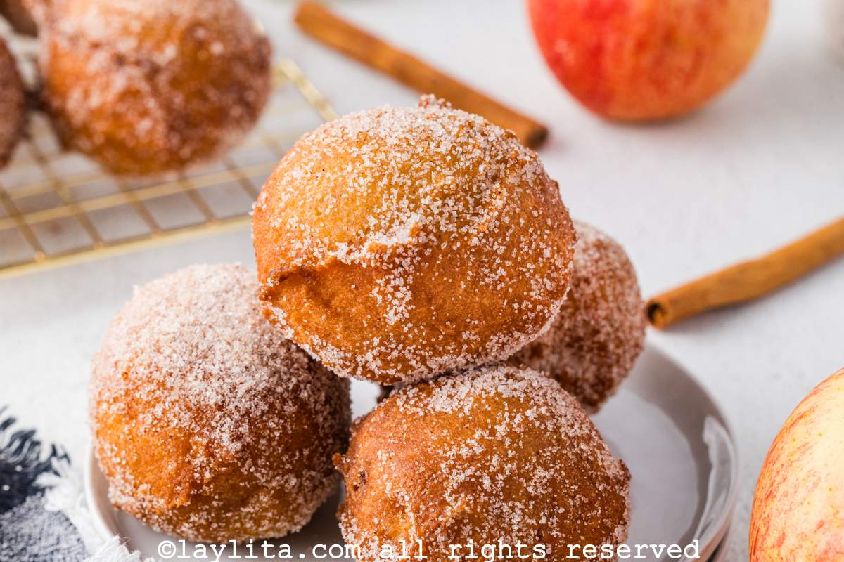 Primer plano de cuatro buñuelos de manzana con azúcar en un plato blanco con astas de canela y una manzana en el fondo