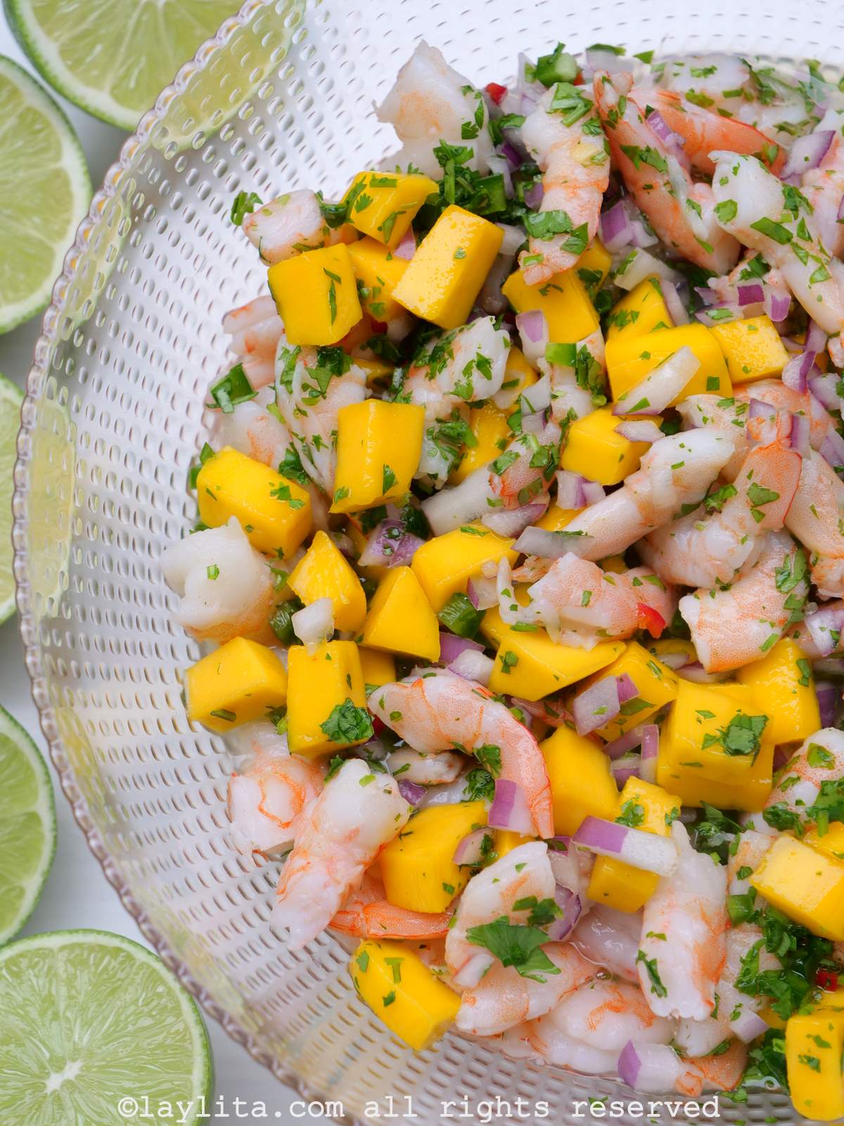 Ceviche de crevettes à la mangue servi dans un bol en verre avec des demi-citrons verts à côté.