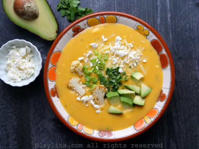 Un bol fleuri rempli de soupe au chou-fleur, garni d'avocat, de fromage et de coriandre, avec une moitié d'avocat en accompagnement.