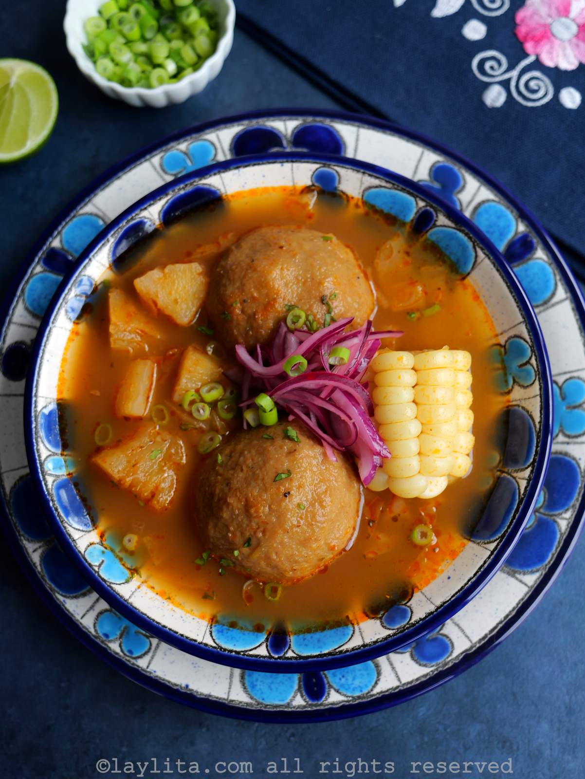 A broth with two green plantains, a piece of corn, other veggies and pickled red onions in a blue floral soup bowl