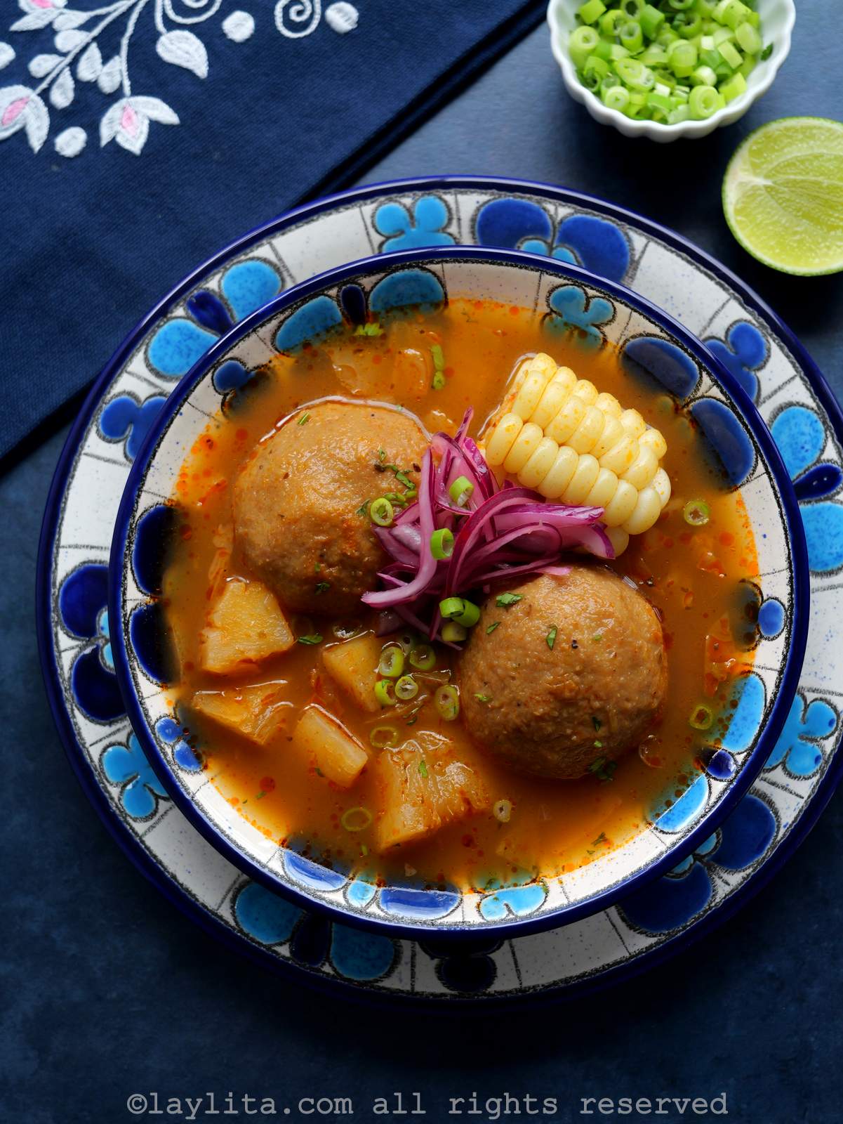 Overhead shot of a broth with two green plantains, a piece of corn, other veggies and pickled red onions served in a blue floral soup bowl