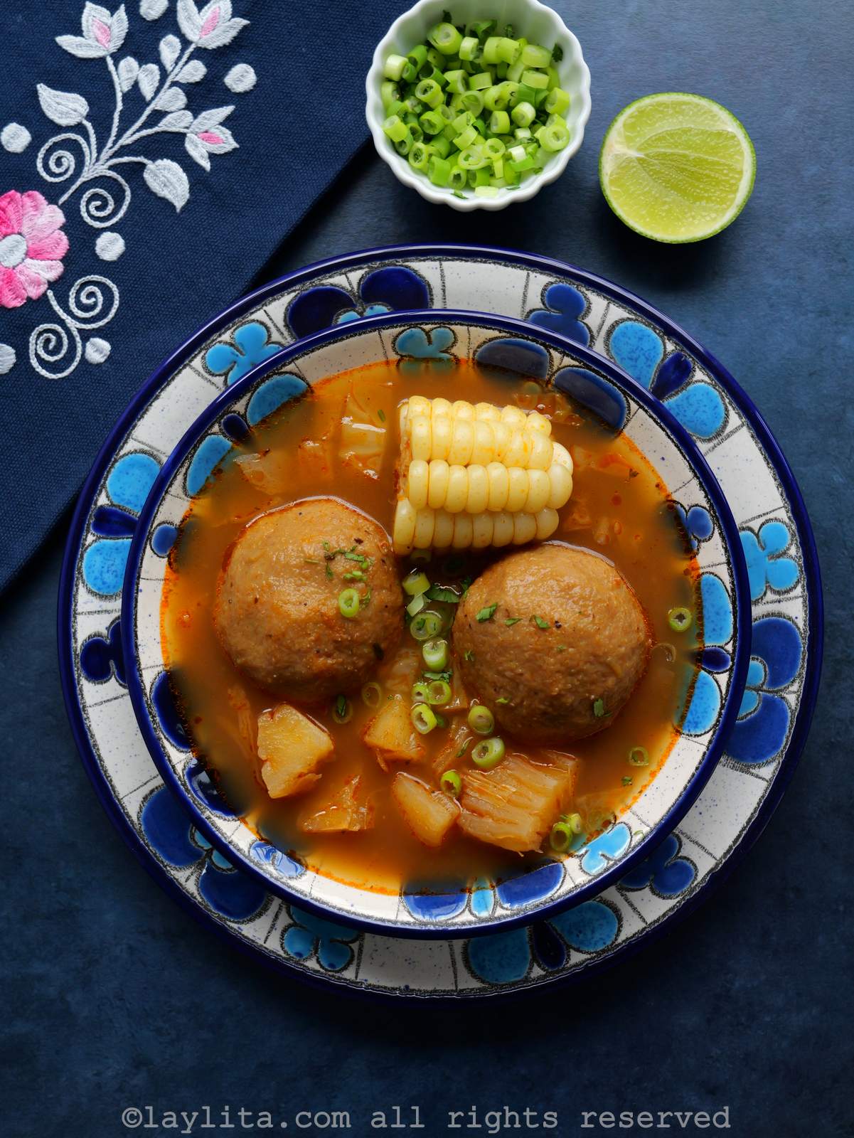 Ecuadorian caldo de bolas served in a blue floral bowl, with a lime half and a small bowl of chopped scallions on the side