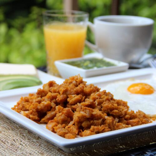 A rectangular white plate with mashed green plantains and a fried egg. A glass of orange juice and coffee cup in the background.