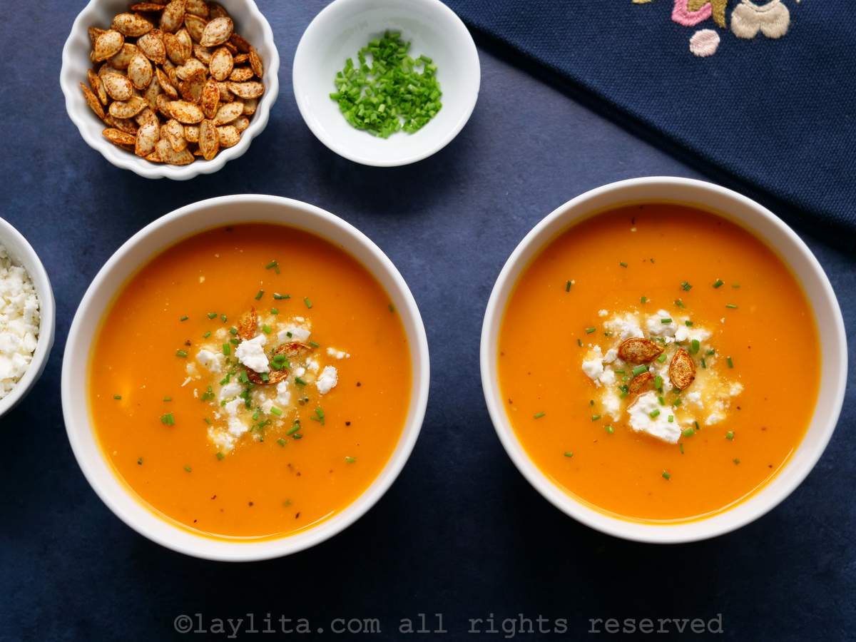 Two bowls of pumpkin or squash soup on a blue background, topped with crispy pumpkin seeds and cheese