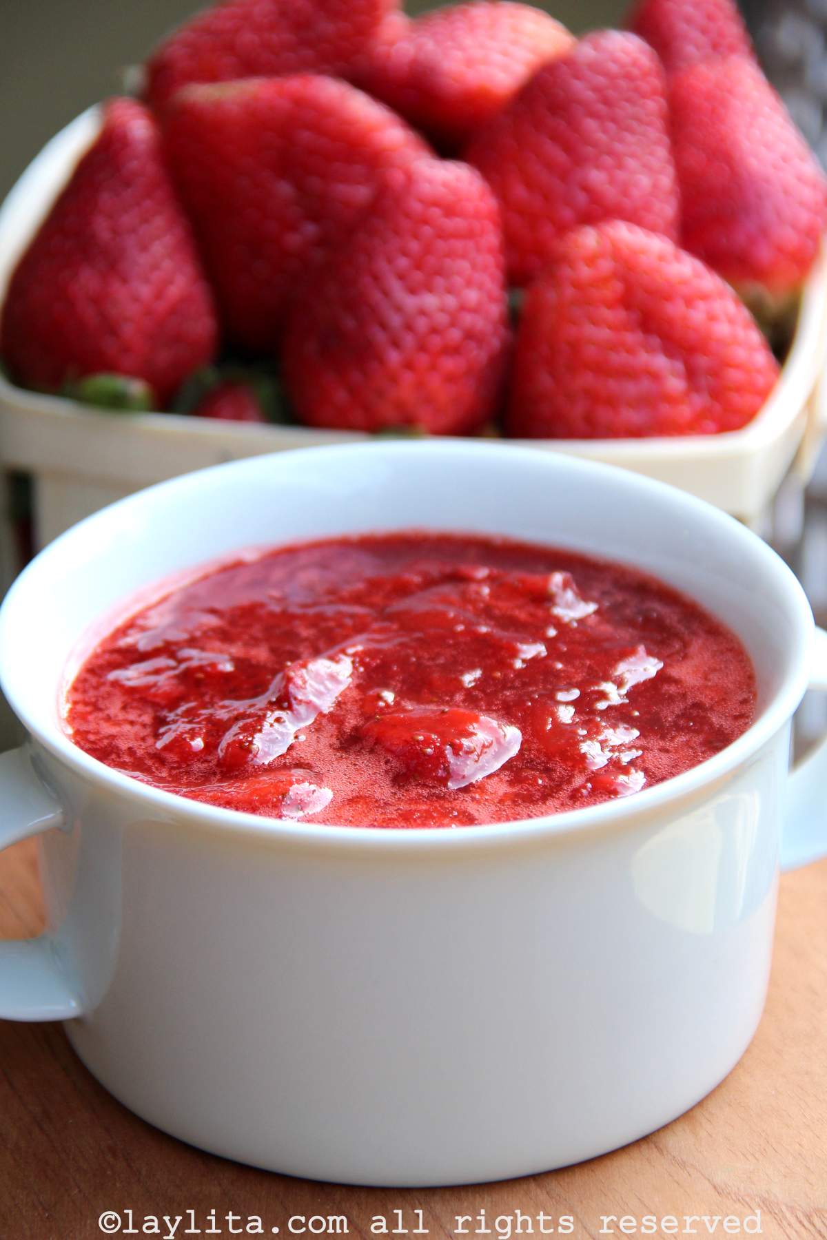 Side view of a bowl of strawberry sauce with a container of fresh strawberries in the background