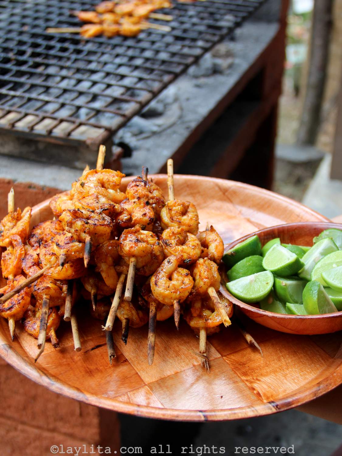 A wood platter with grilled shrimp next to bowl of lime wedges with a grill in the background