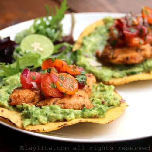 A white plate with two cirspy corn tostadas topped with guacamole, grilled fish, and citrus salsa
