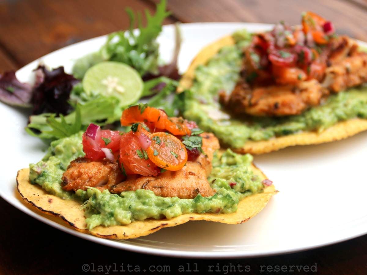 A white plate with two cirspy corn tostadas topped with guacamole, grilled fish, and citrus salsa