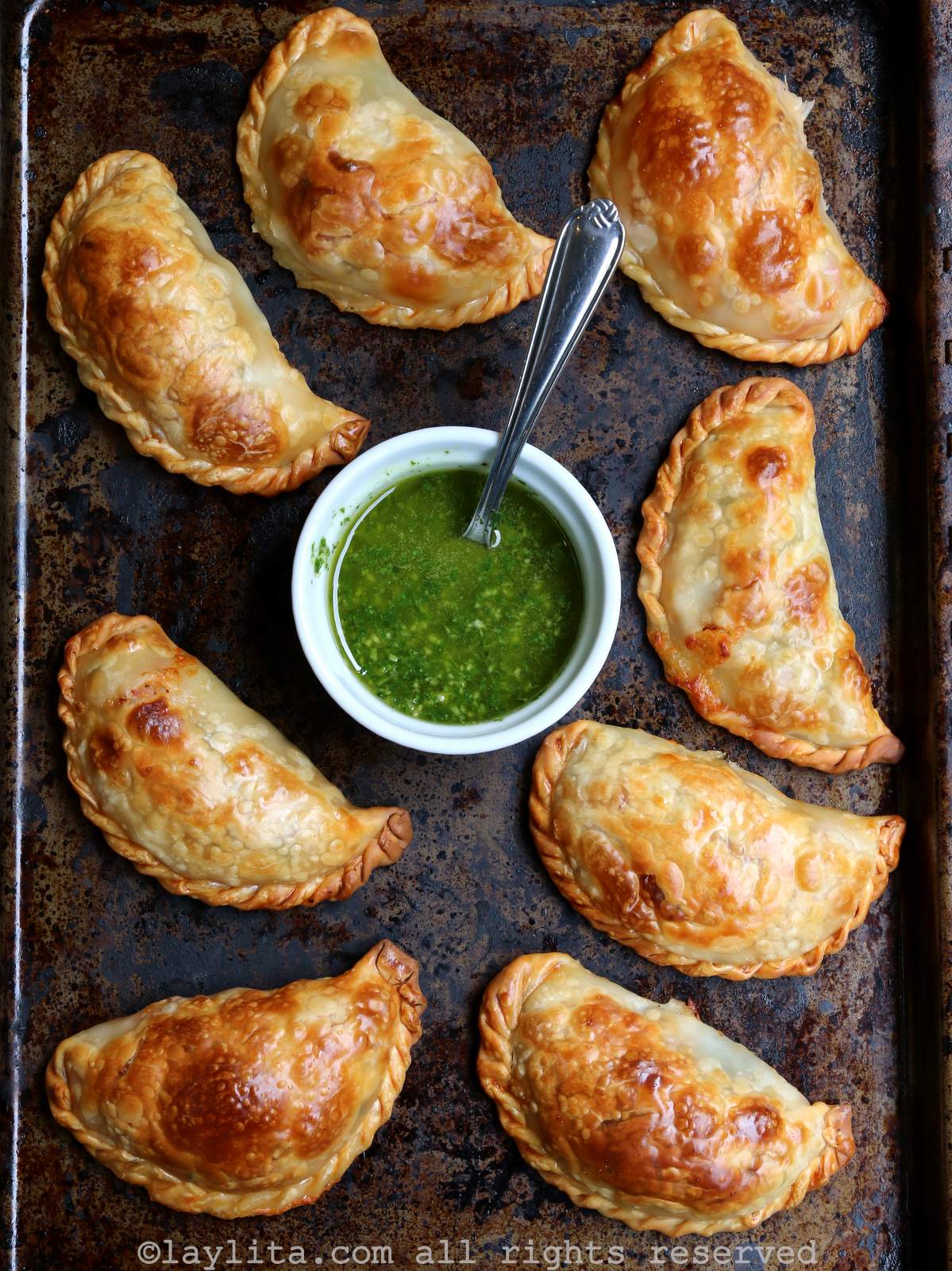 Overhead shot of baked caprese empanadas on a baking sheet with a bowl of green sauce in the middle