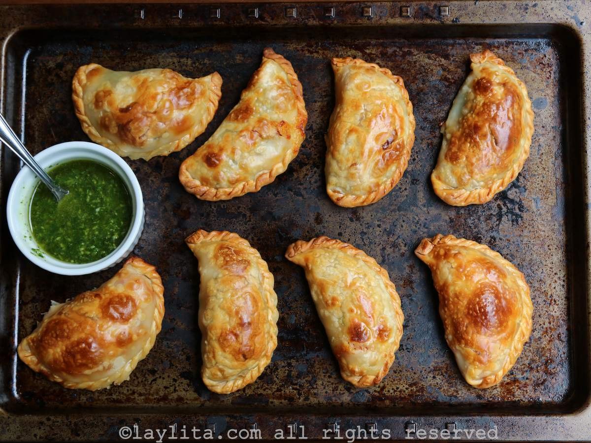 Ten baked empanadas on a baking sheet with a side of green basil dipping sauce