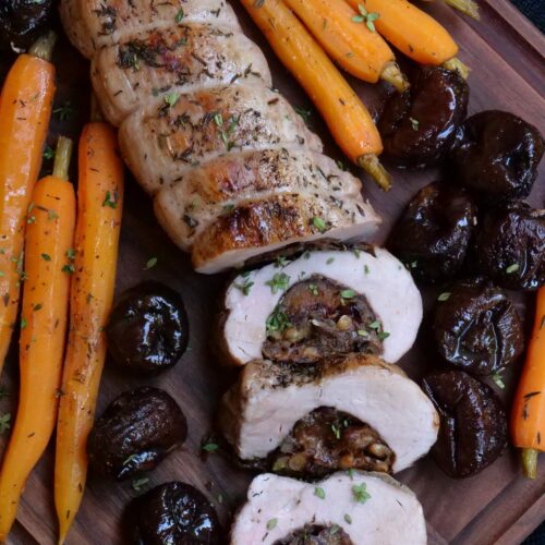 A roasted stuffed pork tenderloin on a cutting board surrounded by roasted carrots and whole prunes