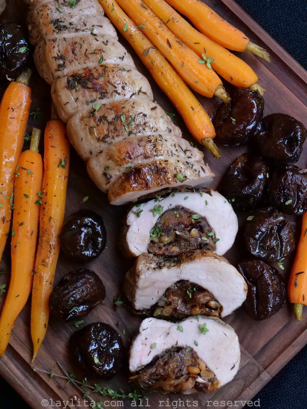 A roasted stuffed pork tenderloin on a cutting board surrounded by roasted carrots and whole prunes