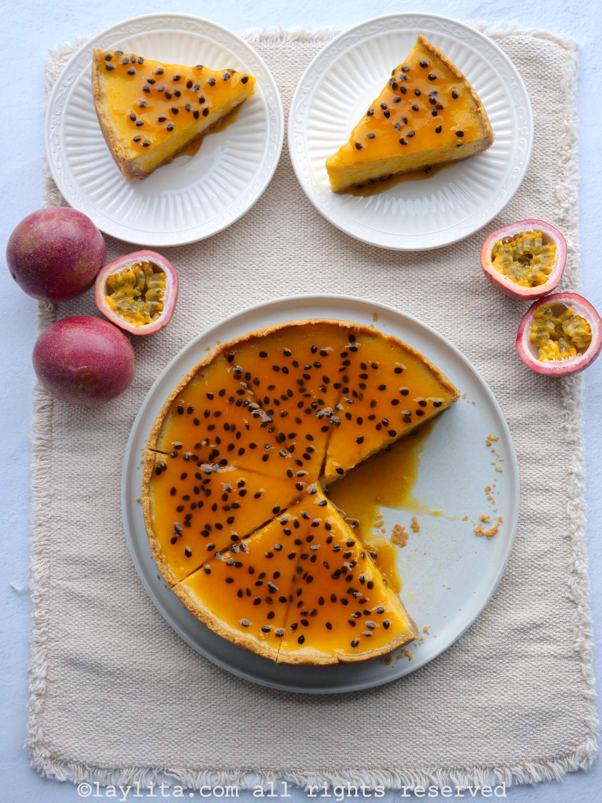 Overhead shot of passion fruit cheesecake and two plates of cheesecake slices on a tan rectangular cloth placemat.